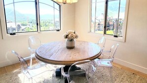 Una mesa redonda de madera con un florero, rodeada de sillas transparentes, en una habitación con ventanas amplias y una lámpara colgante.