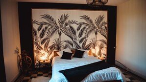 A bedroom with a bed, a headboard with a tropical leaf pattern, a small round table, and a bicycle. - Cocon Bord de Lac, 11 Personnes, Piscine, Billard (PLIEUX)