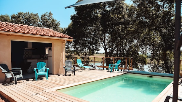 A pool area with wooden decking, lounge chairs, and a small building with a tiled roof. - Cocon Bord de Lac, 11 Personnes, Piscine, Billard (PLIEUX)