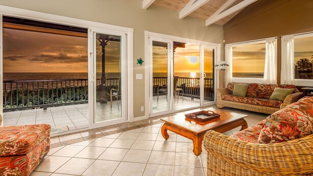 A spacious living area with a view of the sunset over the ocean, featuring a wicker sofa, a wooden coffee table, and a balcony with glass doors.