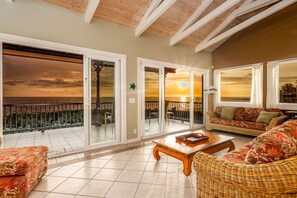 A spacious living area with a view of the sunset over the ocean, featuring a wicker sofa, a wooden coffee table, and a balcony with glass doors. - HOKU'EA HALE  Oceanfront Private Home with Hot Tub (Kailua Kona)
