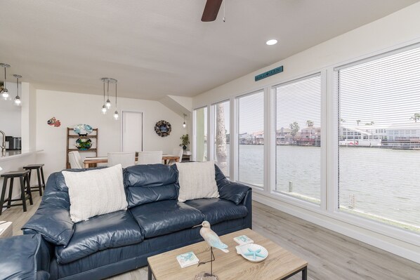 A modern living room with a dark leather sofa, a wooden coffee table, and large windows overlooking a body of water.