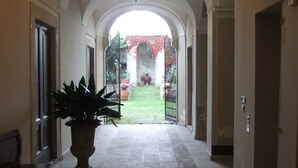 Un couloir avec un plafond haut, un lustre et une vue sur une cour intérieure agrémentée d’une porte et de plantes.