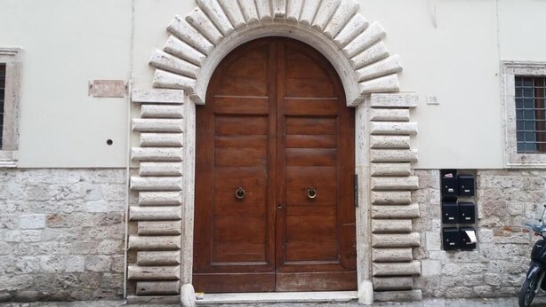 Une grande porte à deux battants en bois, surmontée d’un arc en pierre, intégrée à un bâtiment aux murs de pierre et dotée d’une petite fenêtre sur le côté gauche.