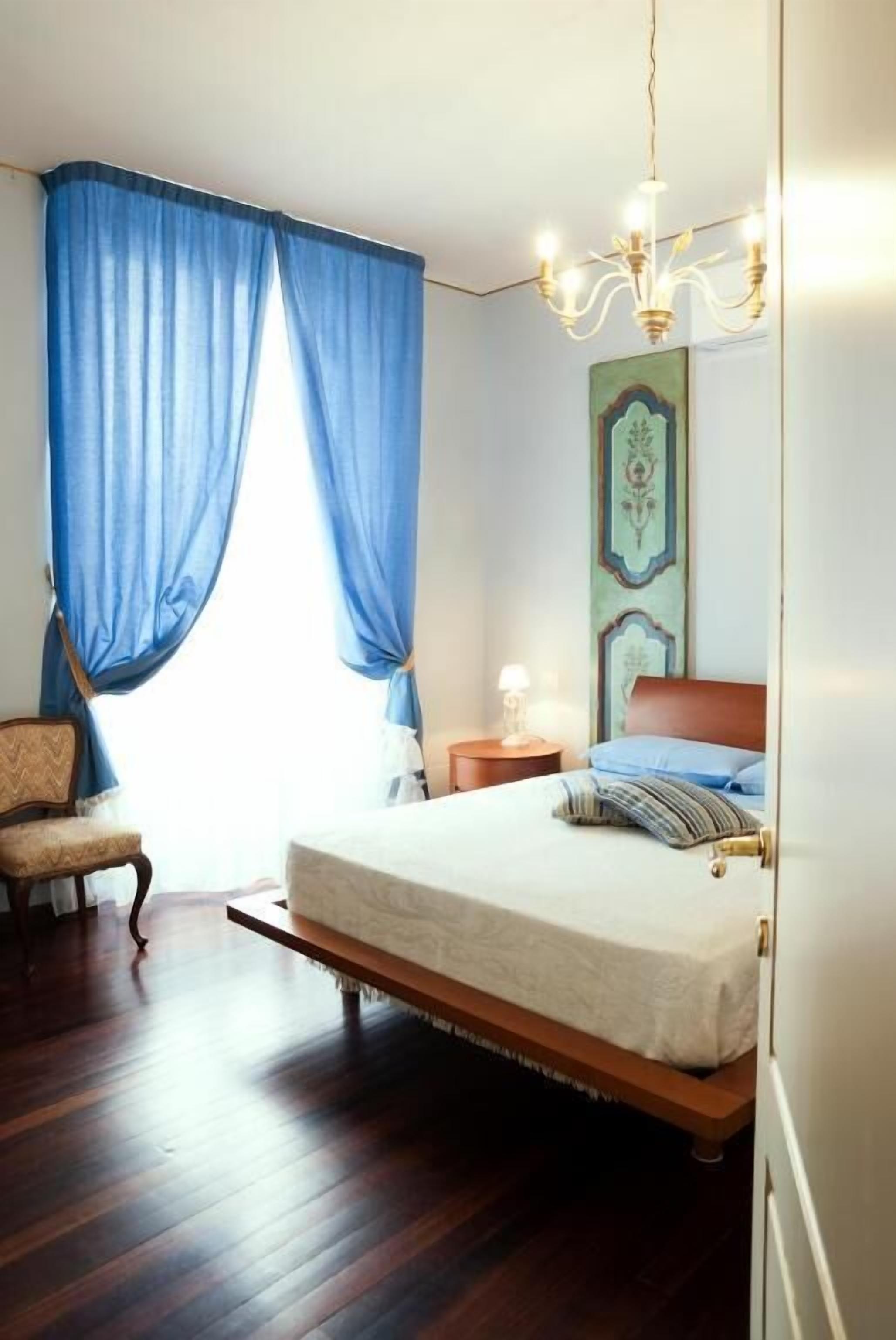 A bedroom with a bed, a chair, a nightstand, a chandelier, and blue curtains.