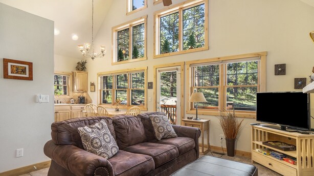 A spacious living room with a brown leather sofa, a black ottoman, and a wooden entertainment center with a flat-screen TV. Large windows provide views of the surrounding trees.