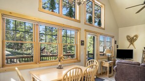 A spacious living room with a wooden dining table, two chairs, a sofa, and large windows offering a view of the outdoors.