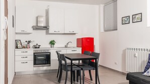 A compact kitchen with white cabinets, a red refrigerator, a built-in oven, and a dining area with a table and chairs.