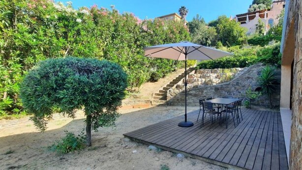 A wooden deck with a table and chairs, surrounded by greenery and a stone wall.
