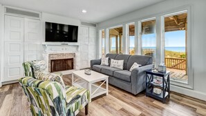 A living room with a fireplace, a sofa, a TV, and large windows overlooking a beach.