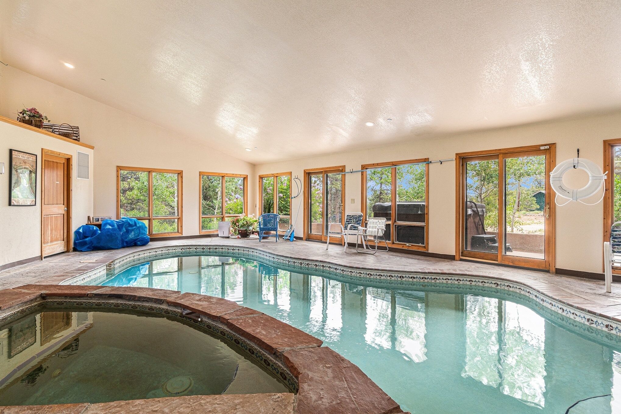 An indoor swimming pool with clear glass walls, a hot tub, and a patio area with chairs and a table.