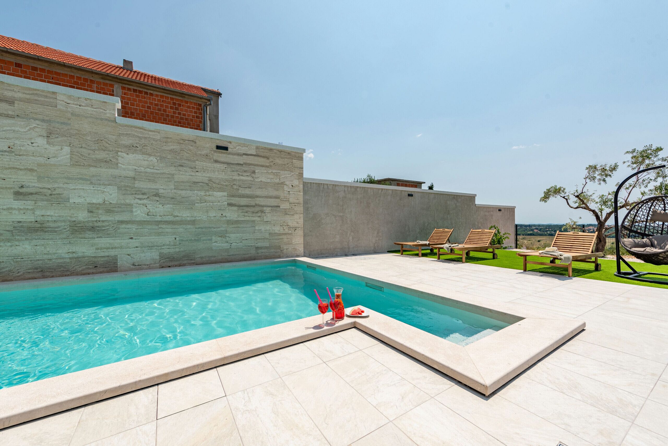 A swimming pool with a tiled deck, a concrete wall, and wooden lounge chairs.