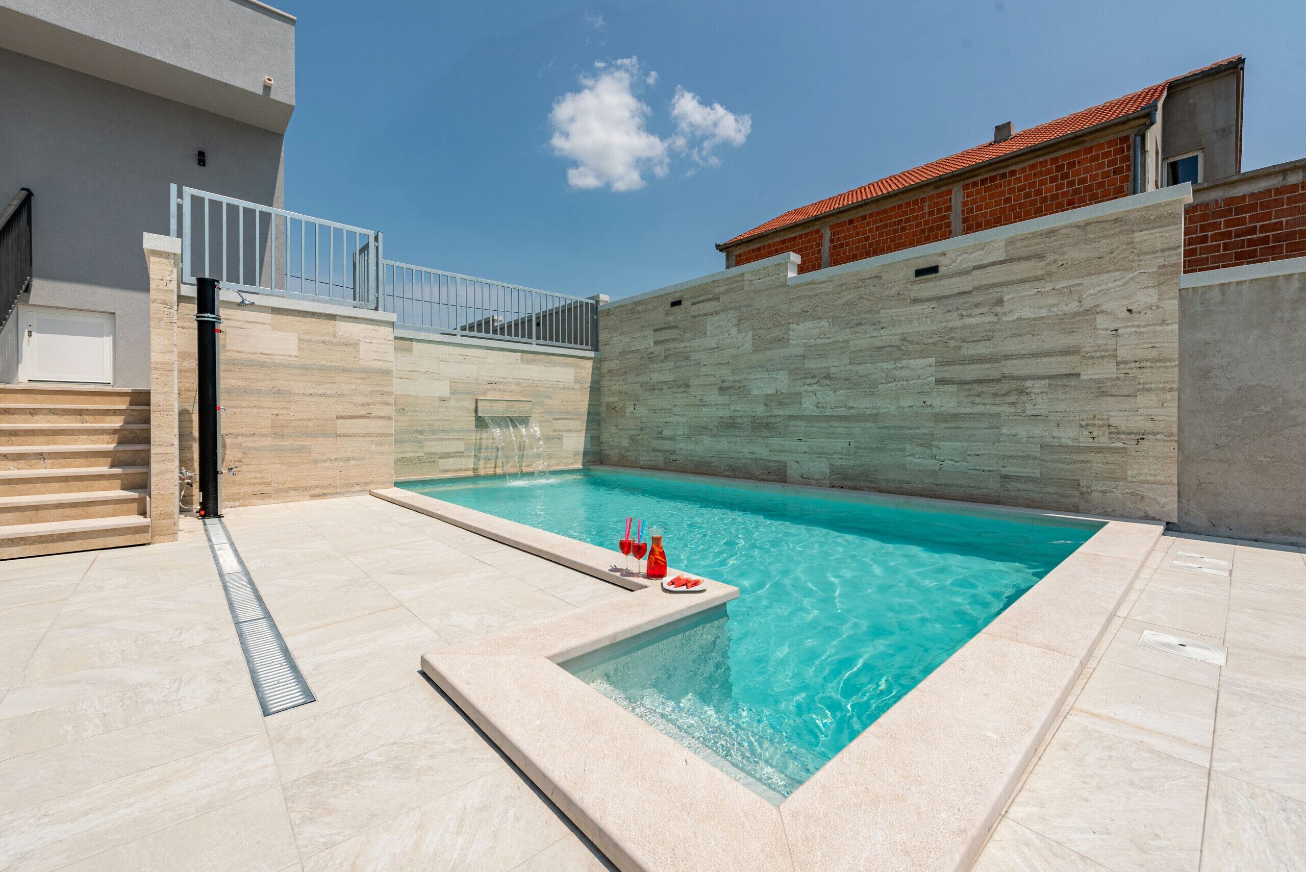 A swimming pool with a stone wall and a metal railing, a tiled deck, and a clear blue sky.