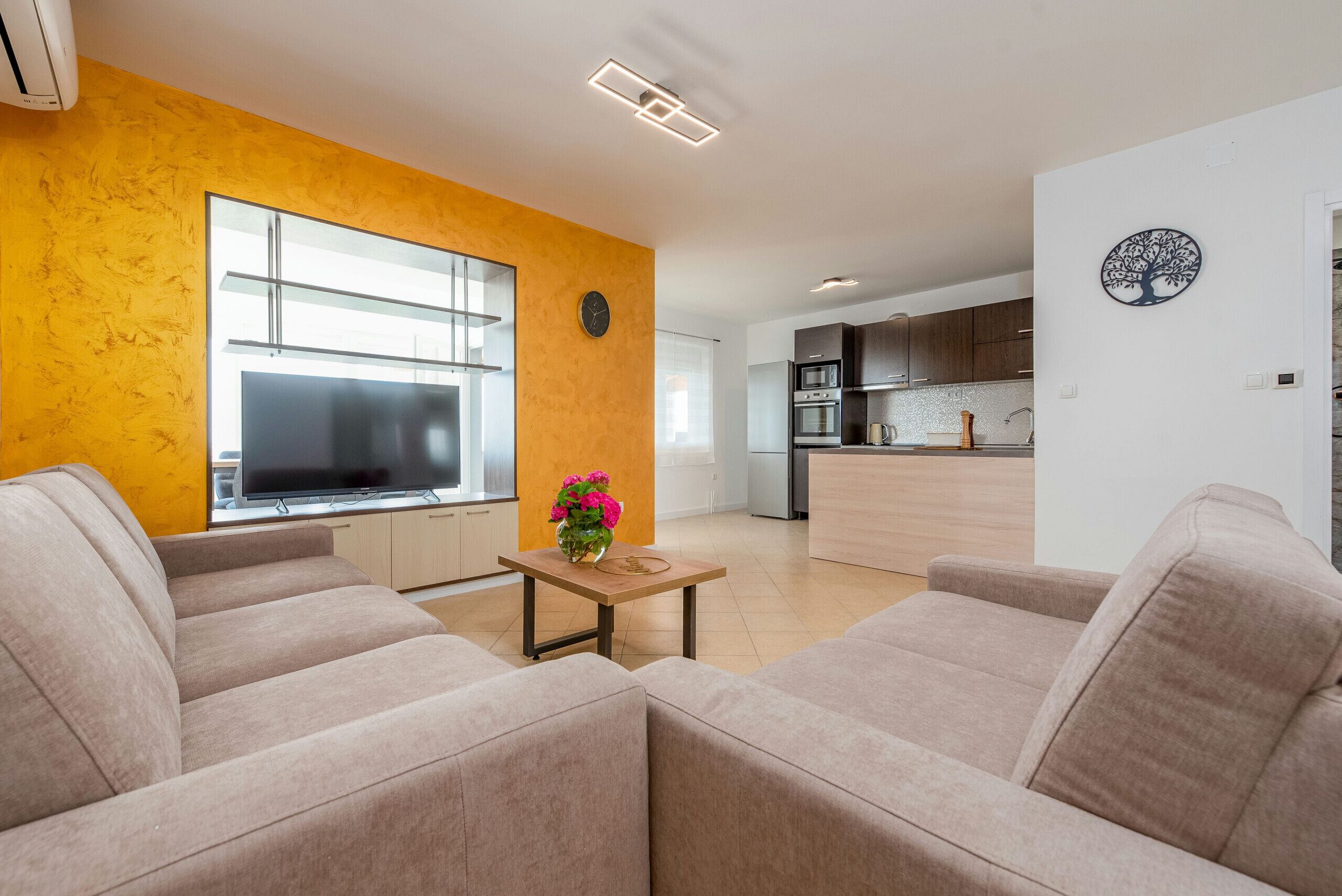 Ein modernes Wohnzimmer mit einer Sofagarnitur, einem Fernseher und einem kleinen Tisch mit Blumen. Im Hintergrund ist eine Küche mit Holzschränken und einer Mikrowelle erkennbar.