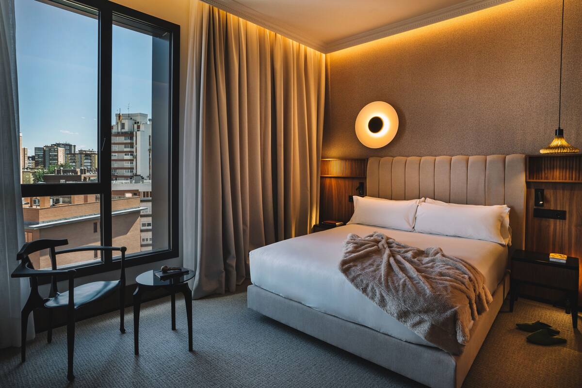 Habitación de hotel con un ventanal grande, una cama con cabecero, una mesita de noche, una silla y una lámpara.
