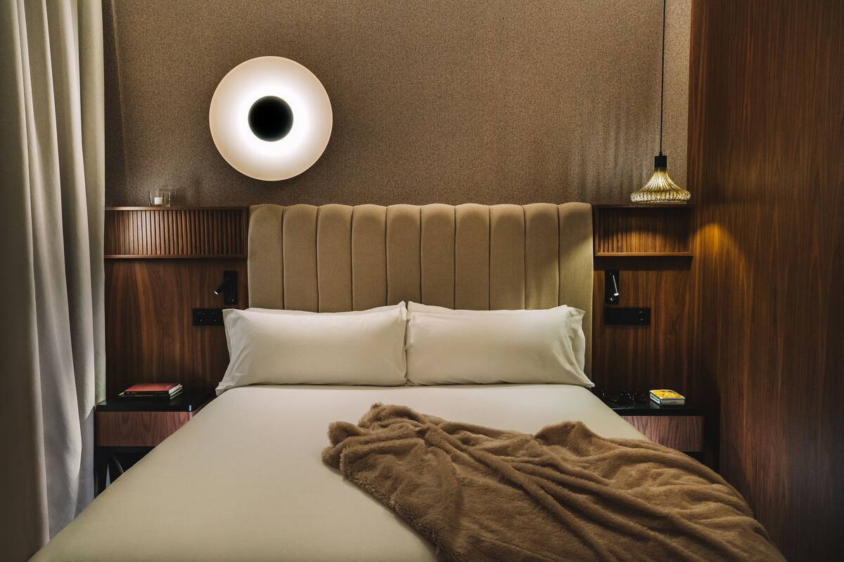 Una cama bien hecha con una manta marrón, una lámpara de pared redonda y una mesita de noche con un libro y una lámpara.