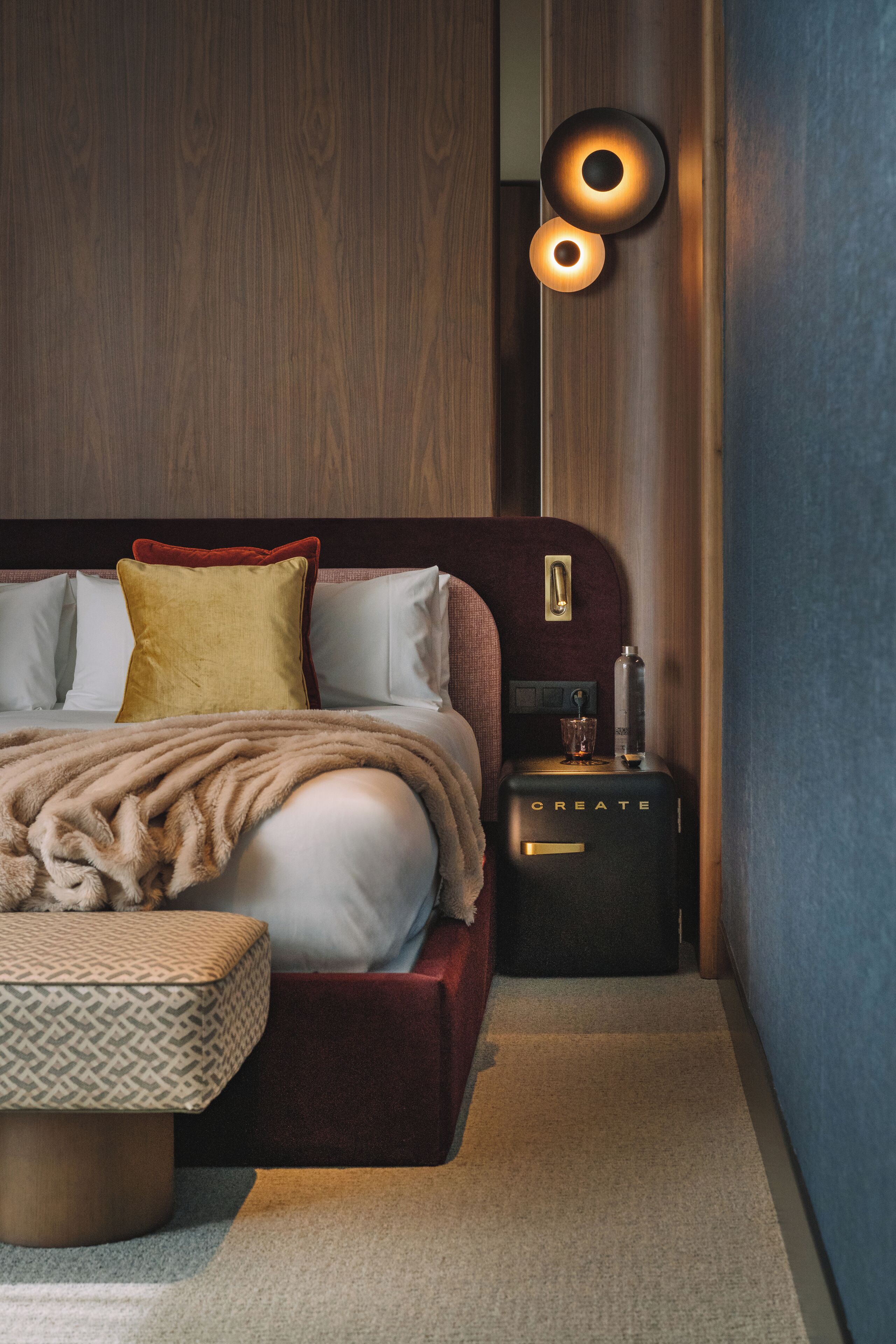 Ein modernes Schlafzimmer mit Holz-Kopfstütze, einem großen Bett mit vielen Kissen, einem Nachttisch mit Flasche und Glas sowie einer Wandleuchte.