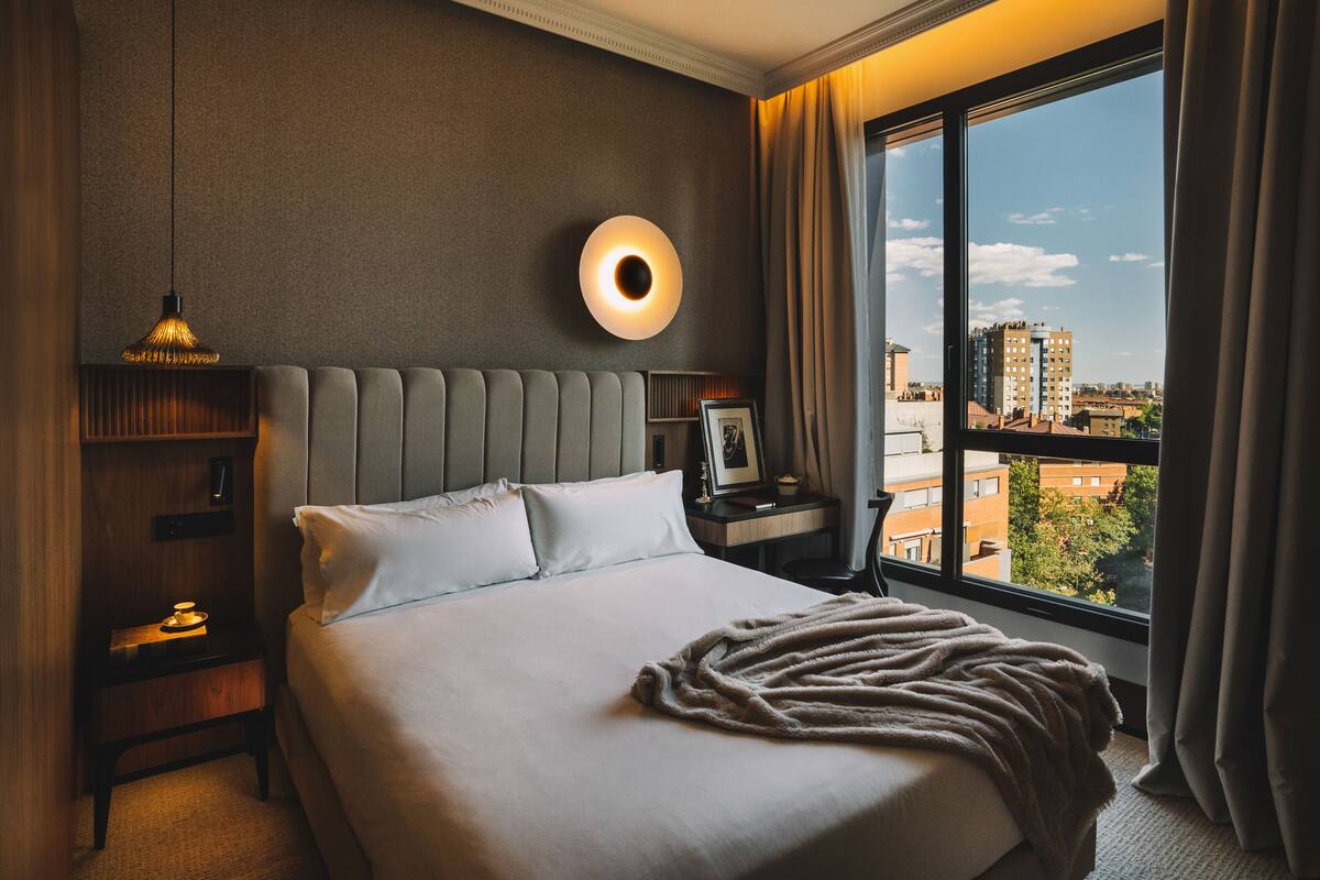 Habitación de hotel con una cama grande, una mesita de noche con lámpara, una luz circular en la pared y vistas a la ciudad a través de la ventana.