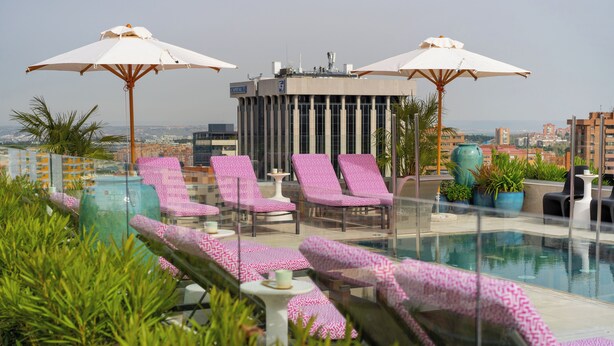 A rooftop pool area with pink patterned lounge chairs, white umbrellas, and a view of the cityscape.