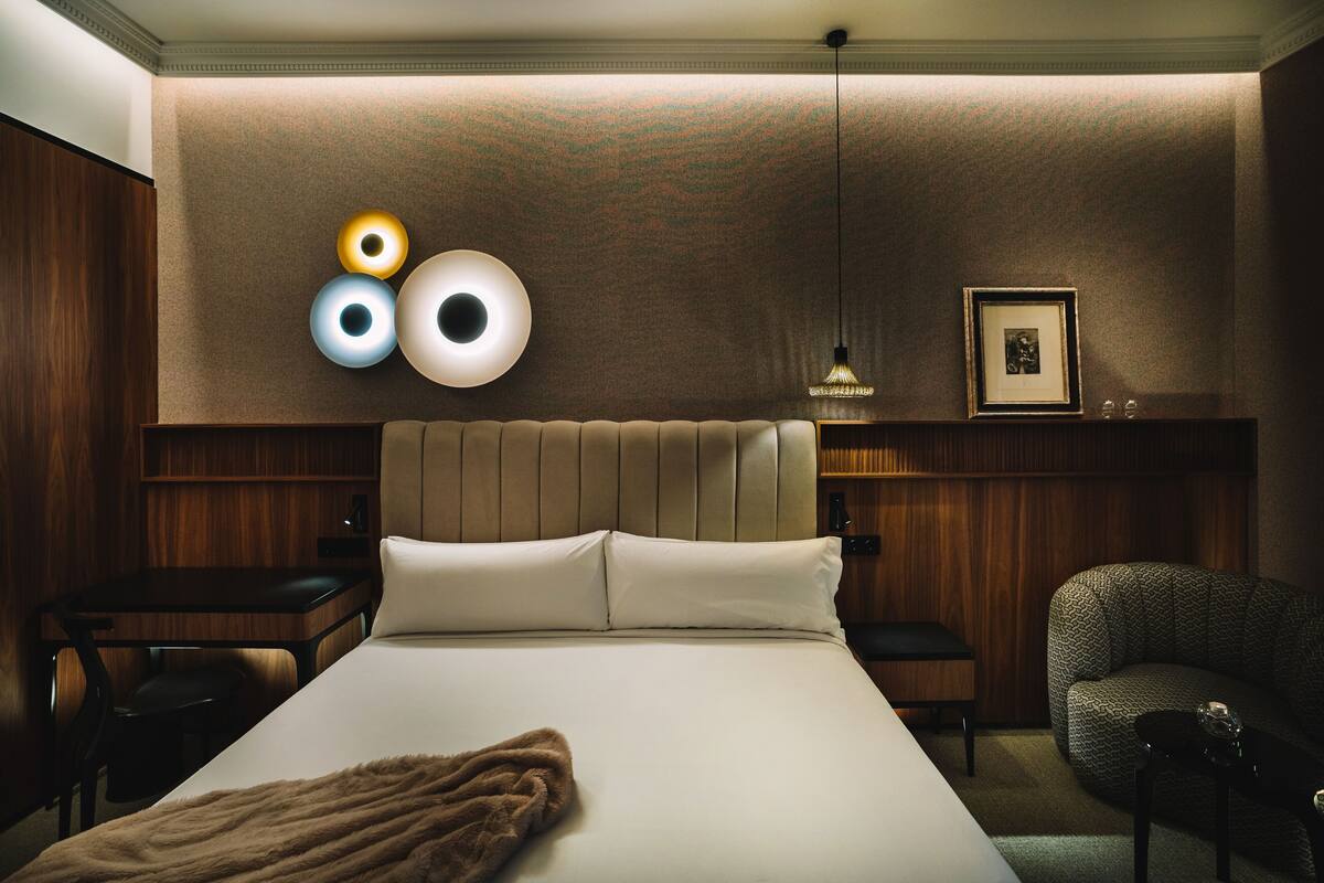 Habitación de hotel con una cama grande, cabecera de madera, mesita de noche, una silla y un aplique de pared.