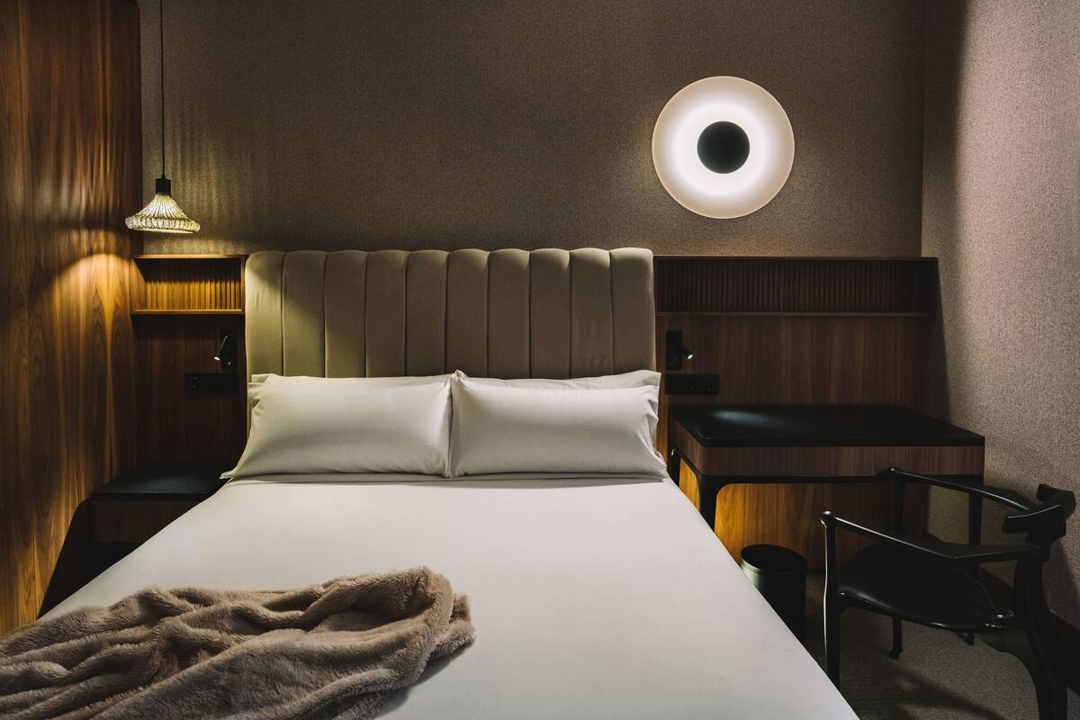 Habitación de hotel con una cama grande, una mesita de noche, una silla, una lámpara de pared redonda y un cabecero de madera.