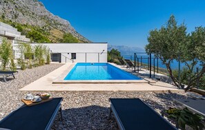Ein Swimmingpool mit Liegestühlen, einer Sonnenterrasse und Blick auf das Meer und die Berge.