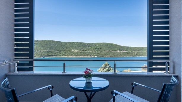 Ein Balkon mit zwei Stühlen, einem kleinen Tisch mit einer Vase und Blick auf eine Küstenlandschaft.