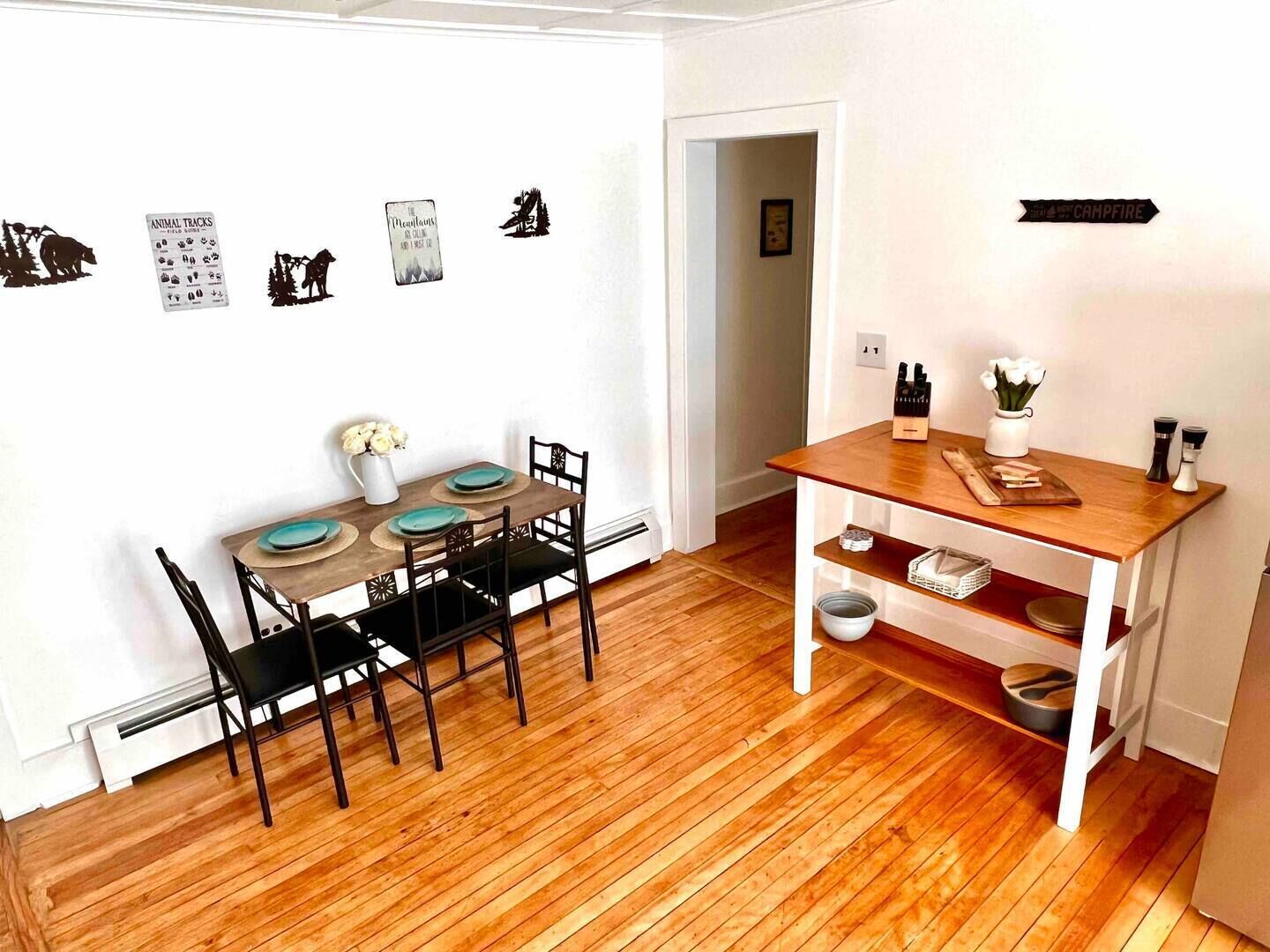 En matplats med ett träbord och stolar, en liten hylla med köksartiklar och väggdekorationer, inklusive djurspår och en bergsbild.
