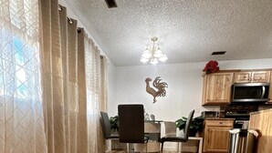 A dining area with a wooden floor, a dining table with chairs, a chandelier, and a kitchen area with cabinets and appliances.