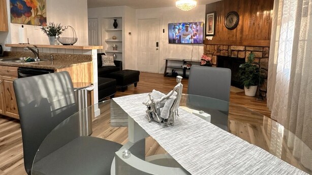 A modern living room with a glass dining table, two grey chairs, a fireplace, a television, and a kitchen area with wooden cabinets and a painting.