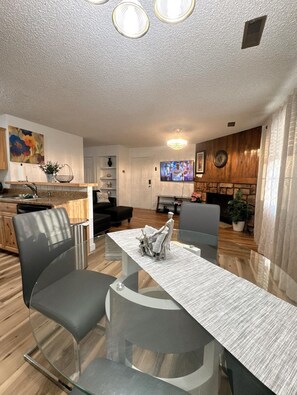 A modern living room with a glass dining table, two grey chairs, a fireplace, a television, and a kitchen area with wooden cabinets and a painting.