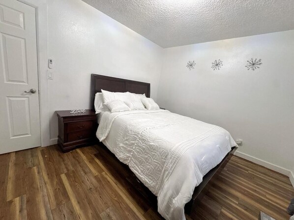 A bedroom with a wooden floor, a bed with white bedding, a nightstand, and a white door.
