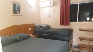 A bedroom with a single bed, a smaller bed, a nightstand, an air conditioner, a window with red curtains, and a small wooden table. - Diroma International Resort - Caldas Novas Goias (Chácara Roma Caldas Novas)
