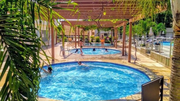A pool area with two pools, covered by wooden pergolas, surrounded by palm trees and lounge chairs. - Diroma International Resort - Caldas Novas Goias (Chácara Roma Caldas Novas)
