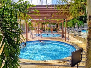 Un área de piscina con dos piscinas, cubiertas por pérgolas de madera, rodeada de palmeras y sillones de descanso.