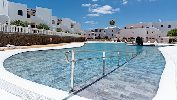 Una piscina con terraza de piedra, edificios blancos al fondo y un cielo azul despejado.