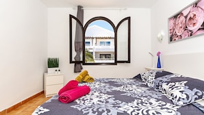 Un dormitorio con una cama grande, una mesita de noche, una planta en maceta y vistas a un edificio a través de una ventana.