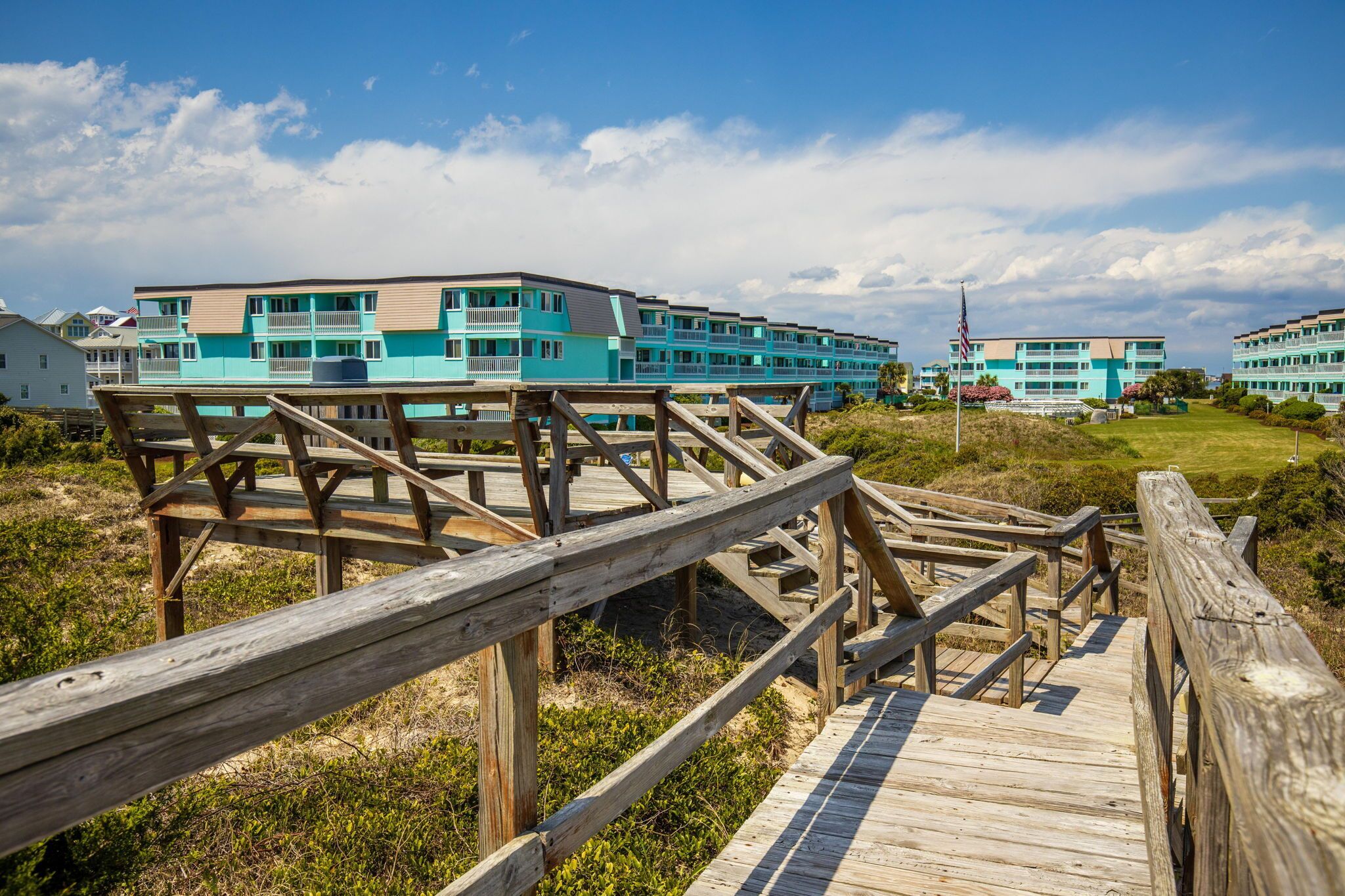 A wooden boardwalk leading to a turquoise hotel with multiple balconies and a clear sky.