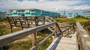 A wooden boardwalk leading to a turquoise hotel with multiple balconies and a clear sky. - Happy Waves-Oceanfront Condo! Sleeps 7! Tons of amenities! (Atlantic Beach)
