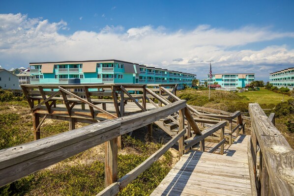 A wooden boardwalk leading to a turquoise hotel with multiple balconies and a clear sky.