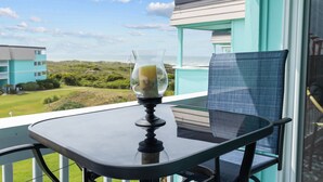 A balcony with a table and chair, a glass pitcher with a beverage, and a view of a building and landscape. - Happy Waves-Oceanfront Condo! Sleeps 7! Tons of amenities! (Atlantic Beach)