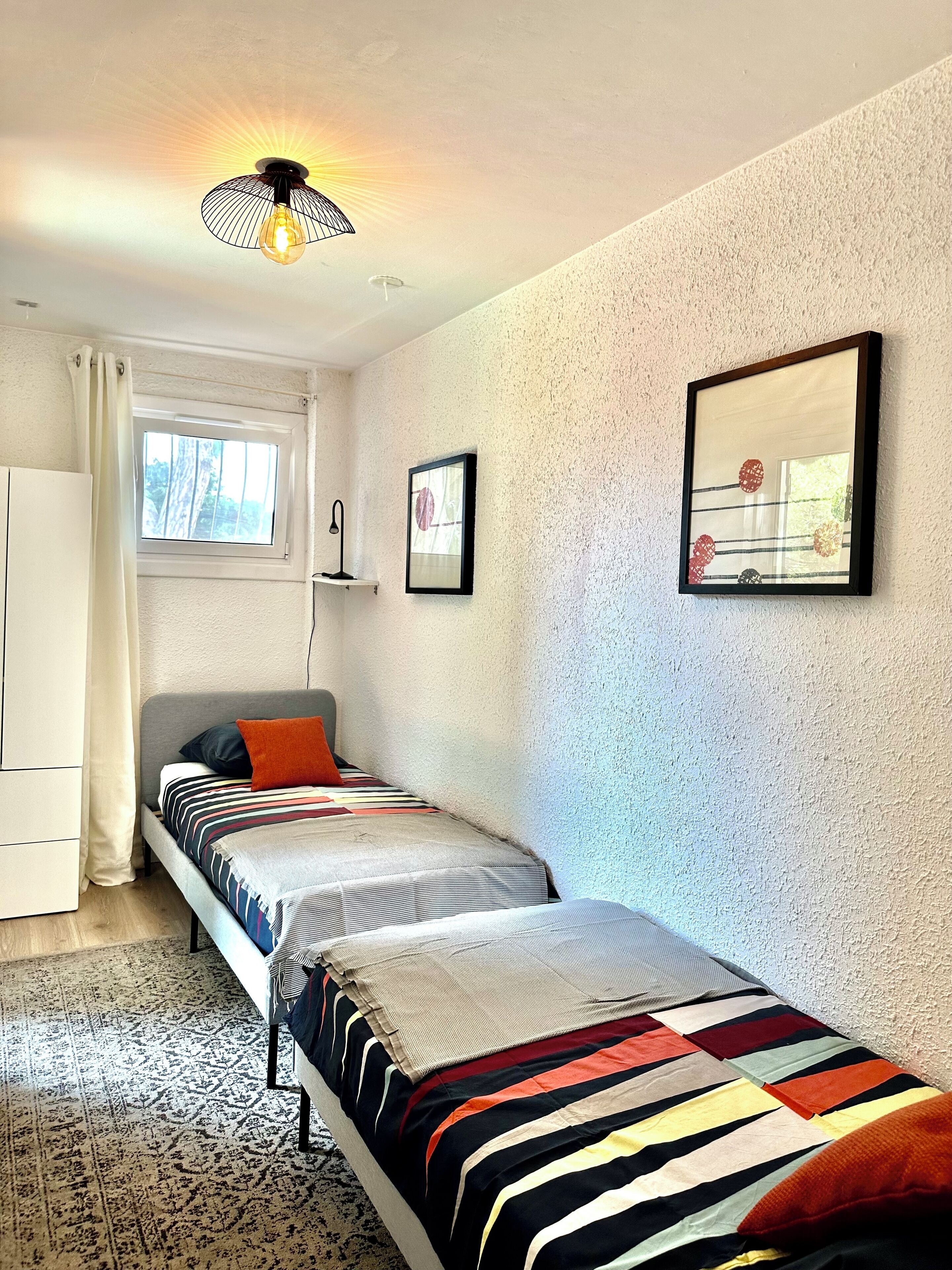 A bedroom with two single beds, a window, a ceiling light, and a framed picture on the wall.