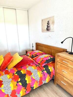 Une chambre à coucher avec un lit, une literie aux couleurs vives, une table de chevet en bois, une lampe et une œuvre d’art murale.