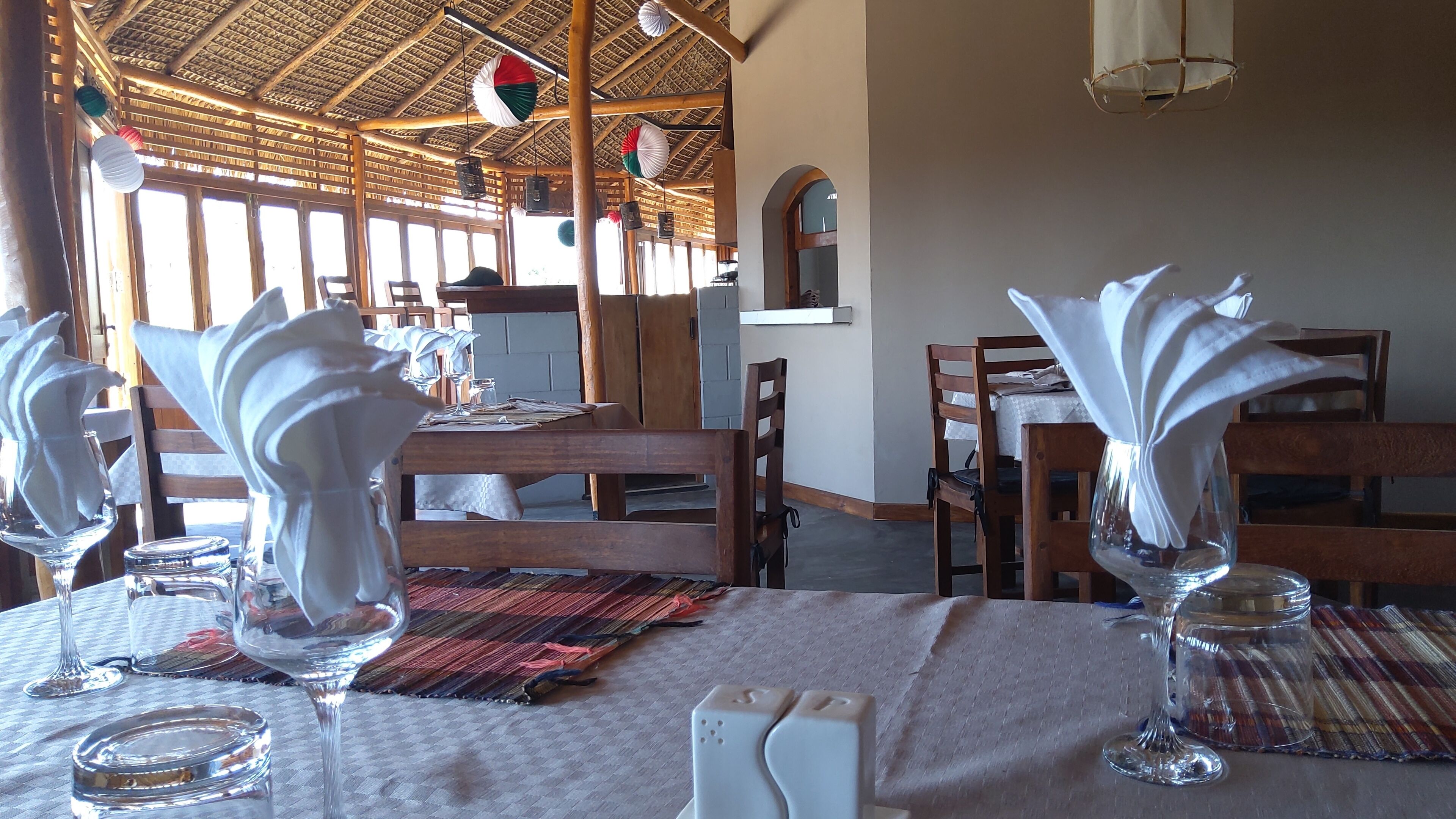 Un restaurante con muebles de madera, mesas dispuestas con servilletas blancas y cubertería de vidrio, y un tejado de paja.