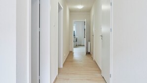 A long, narrow hallway with white walls and wooden flooring, leading to a room with a window and a radiator. - Apartamenty Swinoujscie- Sienkiewicza 23 (Swinoujscie)