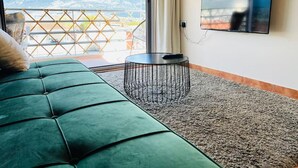 A green sofa, a small round table, and a TV on the wall in a room with a view of mountains through a window. - View Lake Attic in Ioannina (Ioannina)