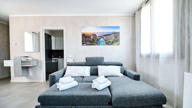 A modern bedroom with a grey sofa bed, a painting of a landscape, a bathroom with a sink, and a mirror. - The Bridge - Peschiera Holiday (Peschiera del Garda)