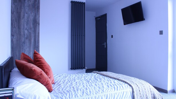 Un dormitorio con cama, almohadas, una mesita de noche, un televisor en la pared, una puerta y un radiador.
