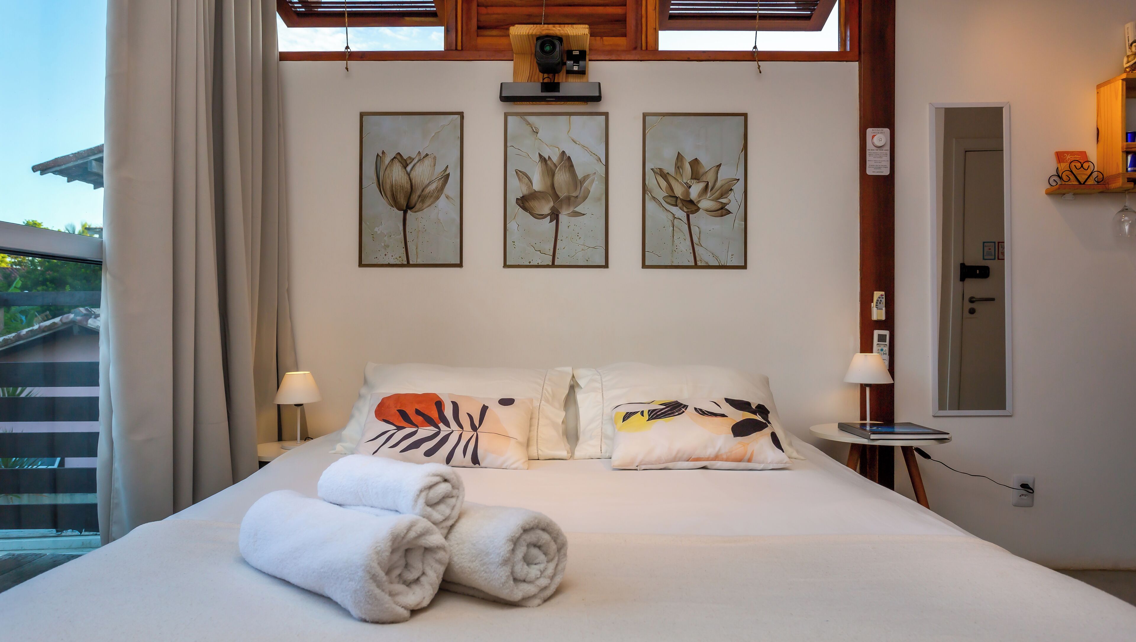 A neatly made bed with towels, three wall-mounted paintings of lotus flowers, a ceiling-mounted camera, and a small bedside table with a lamp.