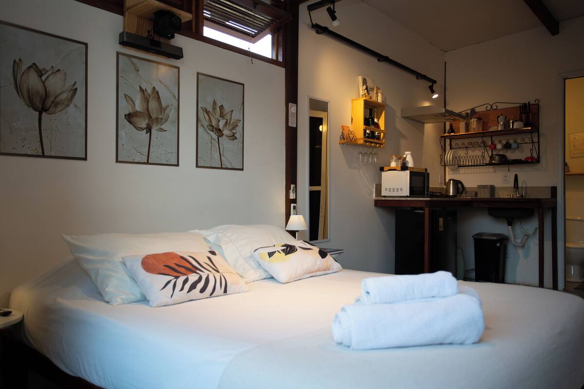 Habitación de hotel con una cama grande, obras de arte en la pared, una zona de cocina y un ventanal que proporciona luz natural.